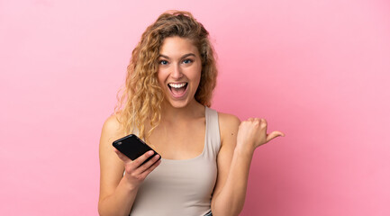 Young blonde woman isolated on pink background using mobile phone and pointing to the lateral