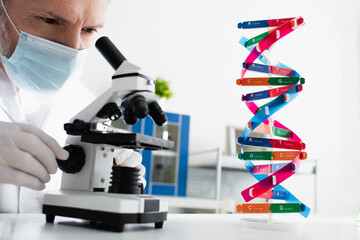 middle aged geneticist in medical mask looking in microscope near dna model.