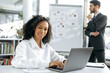 © Kateryna - Successful African American woman sits at her desk in front of a laptop, looks at the camera, smiles. In the background, a male colleague works with charts, develops a strategy for modernizing work