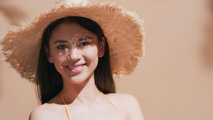 Wall Mural - Close-up beauty portrait of young dark-haired woman in the straw hat hiding from the sun turns to the camera and smiles against a beige background | Sunscreen concept