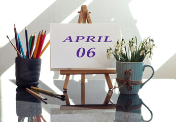 Calendar for the sixth of April: an easel with the inscription April in English and the numbers 03, a bouquet of snowdrops in a cup, multi-colored pencils, brushes in the sun from the window