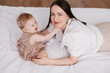 © Tatyana - Sweet young mother playing with her little 1-year-old daughter in bed.Caring brunette woman laying with her baby child at home. Tenderness and love between mother and daughter. Lovely family indoors.
