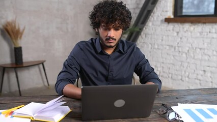 Wall Mural - Portrait of smiling and friendly multi ethnic male freelancer using laptop and looks at the camera, indian latin curly guy in smart casual shirt typing, working remotely, develops software