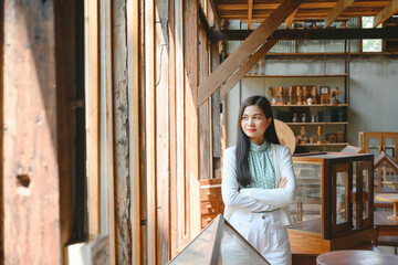 A confident smiling Asian woman stands near her desk at home and looks at the camera in a friendly manner. She owns a piece of handcrafted wood. Work in a wood crafts workshop