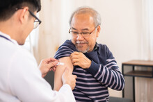 Safe Vaccination For Old People. Elder Man In Medical  Getting Flu Or Covid-19 Vaccine Sitting On Sofa At Home. Asian Doctor Or Nurse Giving Flu Or Covid-19 Shot To Senior.