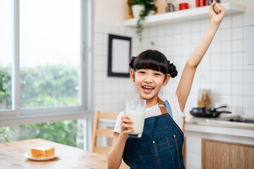 Asian little cute kid holding a cup of milk in kitchen in house. Young preschool child girl  stay home with smiling face, drinking milk and then look at camera.Milk for good health.