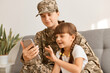 © sementsova321 - Portrait of young adult Caucasian military woman wearing camouflage uniform and cap posing with her daughter, mother holding mobile phone, kid trying to touch phone screen.