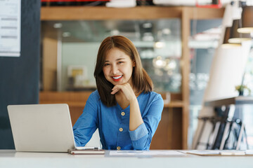 Attractive Asian Young woman freelancer working at the office using a laptop computer Looking at the camera at her comfortable workplace.