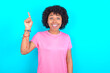 © Roquillo - young girl with afro hairstyle wearing pink T-shirt over blue background pointing finger up and looking inspired by genius thought, showing good idea sign, having clever solution in mind