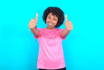 young girl with afro hairstyle wearing pink T-shirt over blue background approving doing positive gesture with hand, thumbs up smiling and happy for success. Winner gesture.