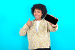 © Roquillo - Portrait young girl with afro hairstyle wearing floral shirt over blue background holding in hands cell showing giving black screen thumb up