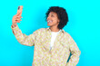© Roquillo - young girl with afro hairstyle wearing floral shirt over blue background smiling and taking a selfie ready to post it on her social media.