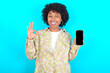 © Roquillo - young girl with afro hairstyle wearing floral shirt over blue background holding in hands cell showing ok-sign