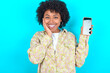 © Roquillo - young girl with afro hairstyle wearing floral shirt over blue background hold hand modern technology use touch face palm astonished impressed scream wow omg unbelievable unexpected