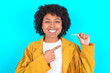© Roquillo - Young woman with afro hairstyle wearing yellow fringe jacket over blue background holding an invisible aligner and pointing at it. Dental healthcare and confidence concept.