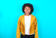 Funny Young woman with afro hairstyle wearing yellow fringe jacket over blue background makes grimace and crosses eyes plays fool has fun alone sticks out tongue.