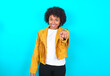 © Roquillo - Excited positive Young woman with afro hairstyle wearing yellow fringe jacket over blue background points index finger directly at you, sees something very funny. Wow, amazing
