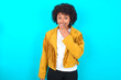 © Roquillo - Young woman with afro hairstyle wearing yellow fringe jacket over blue background covers mouth and looks with wonder at camera, cannot believe unexpected rumors.