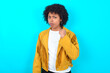 © Roquillo - Young woman with afro hairstyle wearing yellow fringe jacket over blue wall shows fist has annoyed face expression going to revenge or threaten someone makes serious look. I will show you who is boss