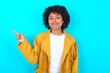 © Roquillo - Young woman with afro hairstyle wearing yellow fringe jacket over blue background points to side on blank space demonstrates advertisement. People and promotion concept