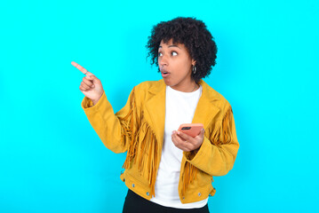 Wall Mural - Stunned Young woman with afro hairstyle wearing yellow fringe jacket over blue background points sideways right copy space, recommends product, sees astonishing thing