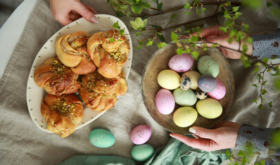 Christian holiday, Easter. Easter buns with pistachios on a white plate, colored eggs with hands on the table. Tree branches, vases. Rustic. Background image, copy space. Top view