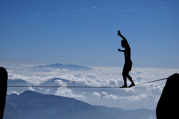 Silhouette of young man balancing on slackline high above clouds and mountains. Slackliner balancing on tightrope beautiful colorful sky and clouds behind, highline silhouette.