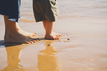 Wall Mural - Baby's first steps. Mother and small child walking barefoot on beach sand near by ocean. Happy young family in nature. Mother and baby playing outdoors on sea beach. Close-up human feet.