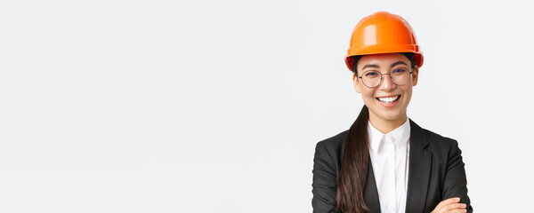 Smiling leader of the team, asian female chief engineer in safety helmet and suit cross arms confident, smiling happy at camera, introduce enterprise, standing white background