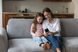 © fizkes - Happy sweet little daughter kid and cheerful mom taking selfie on smartphone, using cellphone on comfortable couch at home, talking on video call. Loving mother and little girl enjoying family leisure
