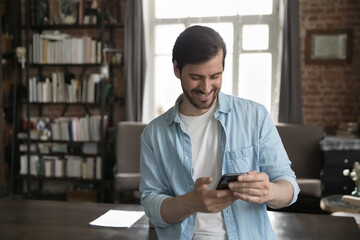 Wall Mural - happy joyful millennial business man using online app on smartphone, standing in home office loft in