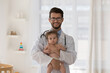 © fizkes - Happy young pediatrician man wearing white coat, holding baby patient in arms, looking at camera, smiling. Kids doctor with few month child head shot professional portrait. Infant health care