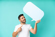 © luismolinero - Young handsome caucasian man isolated on blue background holding an empty speech bubble and pointing it