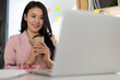 © Wasan - Charming Asian businesswoman in office smiling and drinking hot coffee while working on laptop.