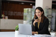 © Wasan - Smiling Asian businesswoman working on laptop and reading documents in office.