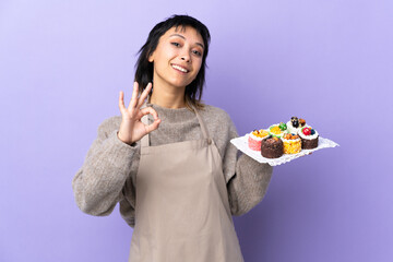 Wall Mural - Young Uruguayan woman holding lots of different mini cakes over isolated purple background showing an ok sign with fingers