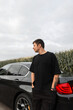© alones - Young businessman in a black T-shirt with a tattoo on arm standing next to a black car on the road outside the city. Travel by car