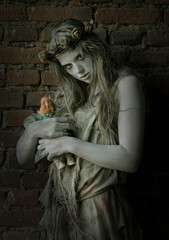 A girl is dressed in rags as a Halloween horror 
figure. She stands in front of an old brick wall. 
Her clothes are torn and dirty and she holds a 
broken doll in her arms.
