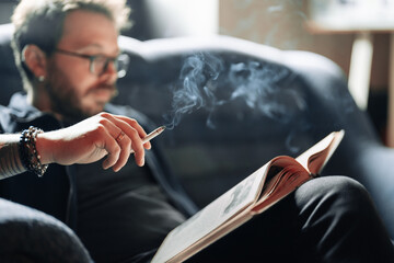 Adult student reading a book in the college library while smoking. Young male wearing glasses with bookshelves on background sitting on couch.