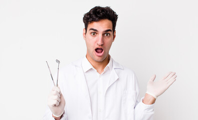 Wall Mural - handsome hispanic man looking surprised and shocked, with jaw dropped holding an object. dentist concept