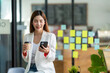 © crizzystudio - Charming Asian woman with a smile standing holding papers and mobile phone at the office.