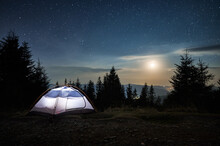 Illuminated Tent Setting Up On Edge Of Stone Trail On Mountain Hill. Incredible Beauty Of Evening Landscape. Moonlight, Low Clouds On Background Of Starry Sky. Mountain Hills In The Distance.