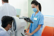 © DragonImages - Serious young medical nurse looking at screen of refractometer machine to check eyes of patient