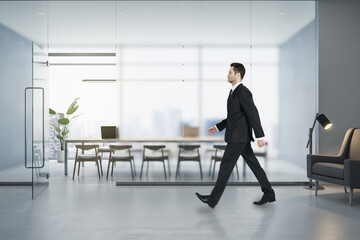 Poster - Attractive young european businessman walking in modern concrete office interior with matte partition glass, furniture, equipment and city view with daylight. Worker and ceo concept.