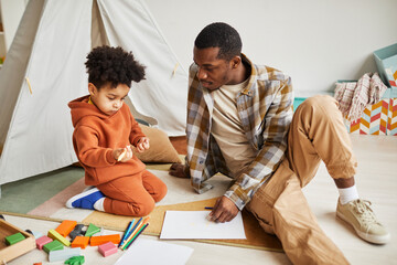 Wall Mural - Full length portrait of caring young father playing with cute toddler boy sitting on floor in cozy kids room, fatherhood and family