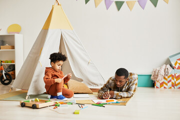 Wall Mural - Portrait of young black father and son playing together on floor in minimal kids room interior with tent and pastel decor, copy space