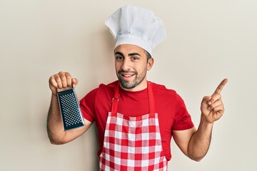 Wall Mural - Young hispanic man wearing professional cook uniform holding grater smiling happy pointing with hand and finger to the side