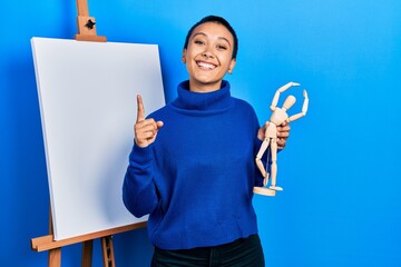 Sticker - Beautiful hispanic woman with short hair holding manikin close to easel stand smiling with an idea or question pointing finger with happy face, number one