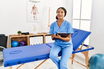 Wall Mural - Young latin woman wearing physiotherapist uniform writing on clipboard at clinic