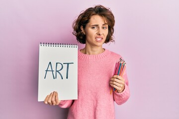 Poster - Young brunette woman holding art notebook and colored pencils clueless and confused expression. doubt concept.